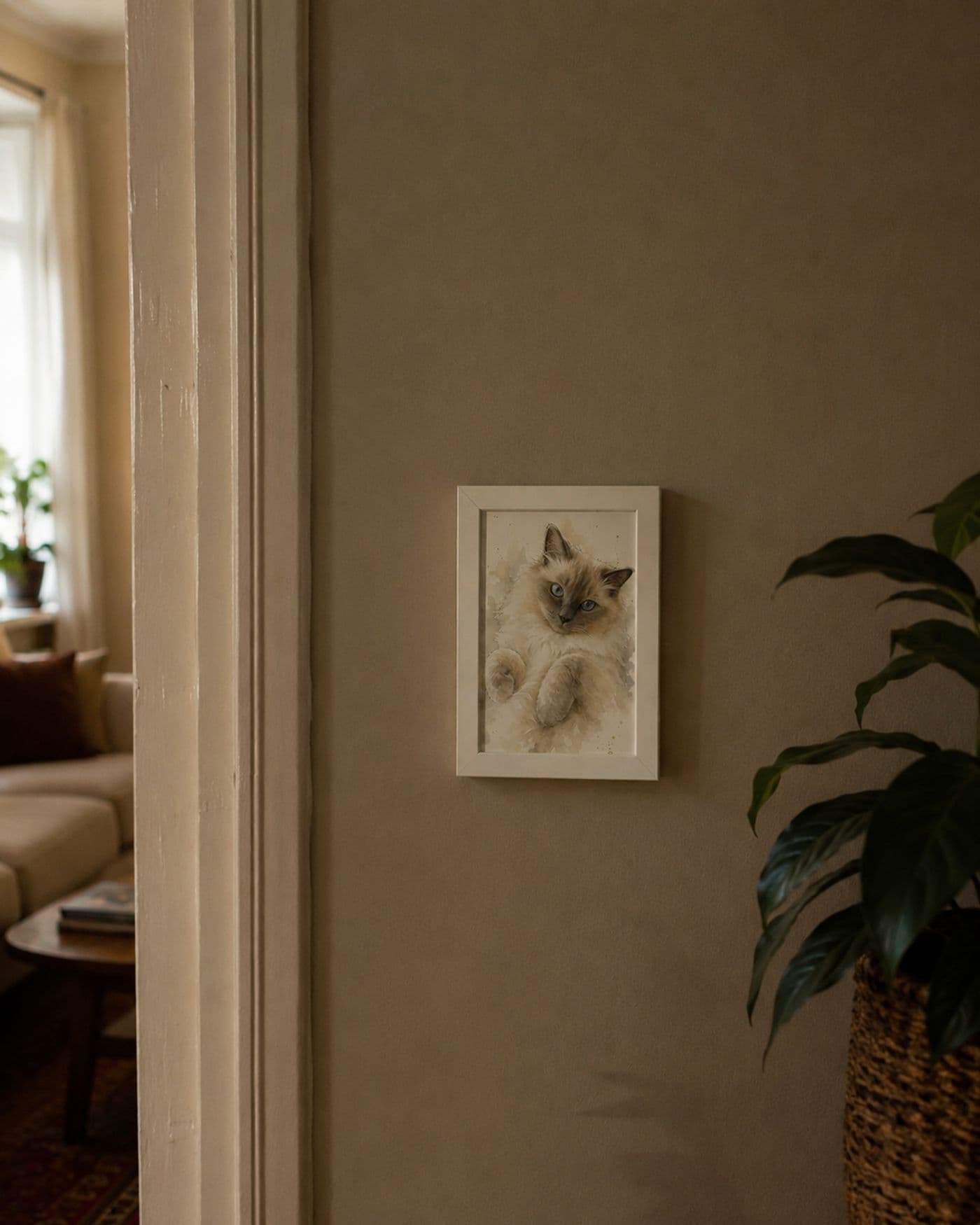 Klein wit ingelijst aquarelportret van een ragdoll aan een warme muur, met doorkijk naar de woonkamer