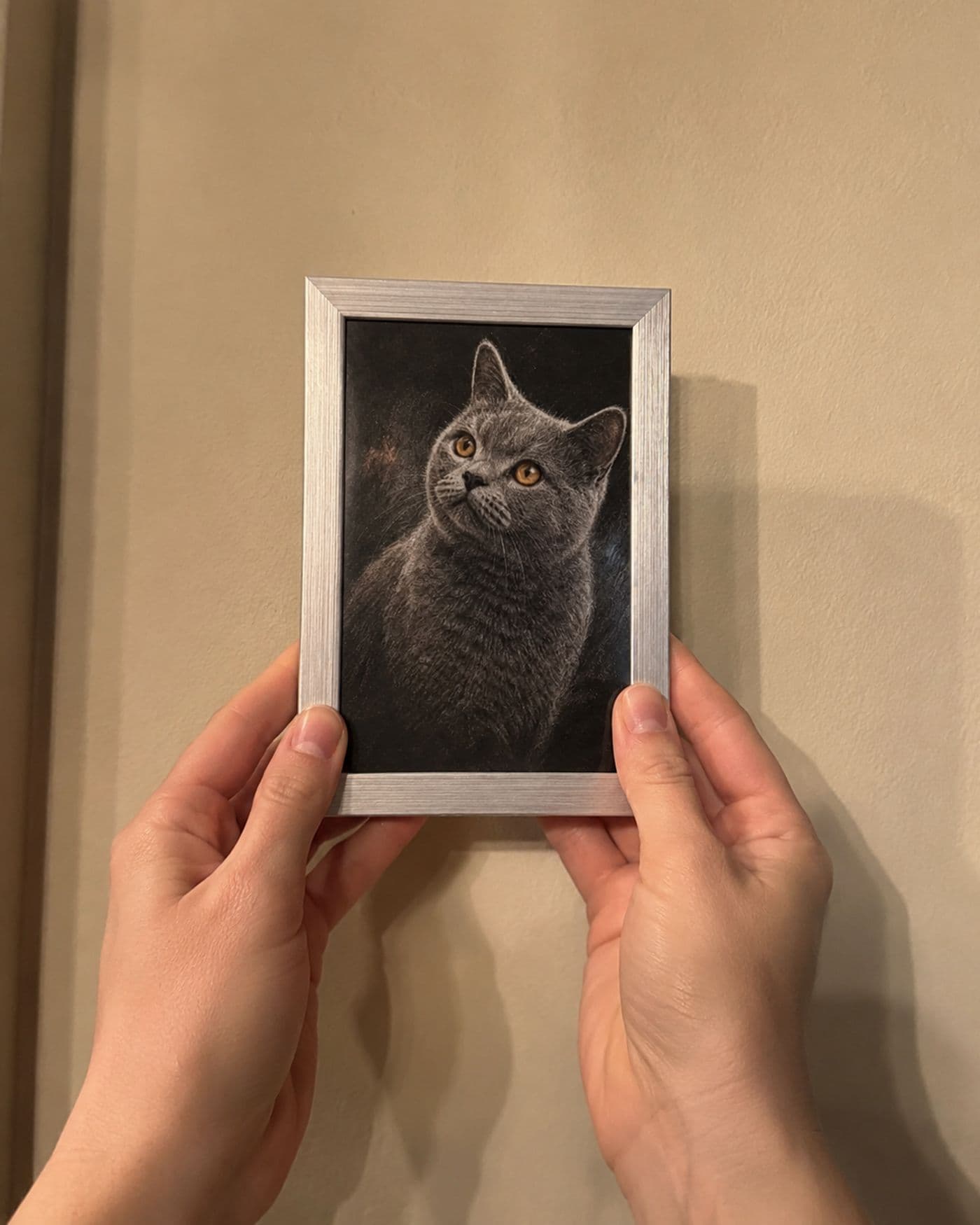 Twee handen houden een klein zilverkleurig ingelijst kattenportret tegen een warme muur, vlak voor het ophangen