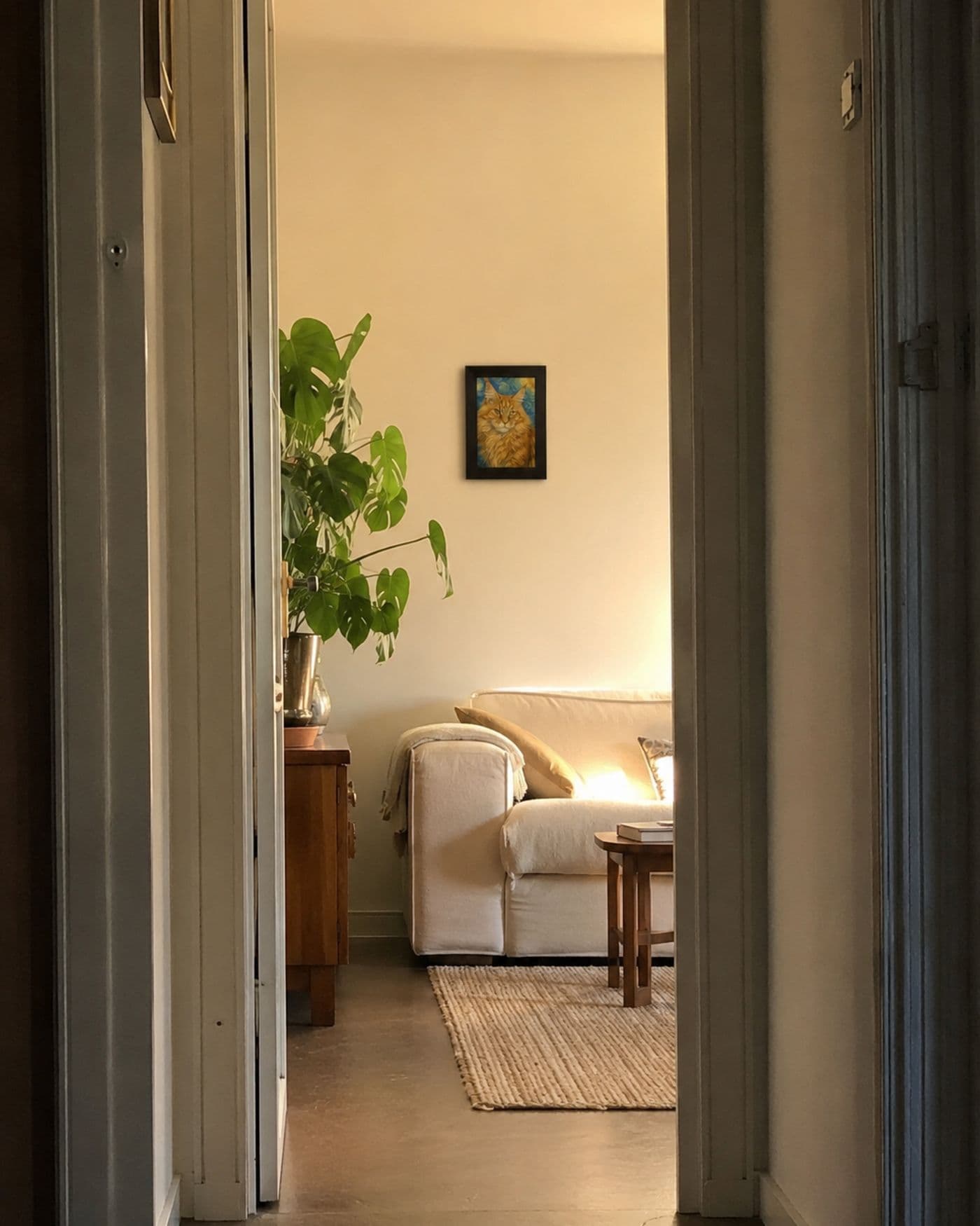Door een openstaande deur zicht op een warme woonkamer met een klein ingelijst kattenportret aan de muur boven de bank